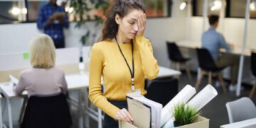 Frustrated young woman in yellow sweater standing at table and touching face with hand while packing stuff in office after dismissal