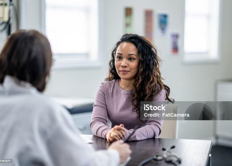 A young woman of mixed race, sits with her female doctor as they discuss her health concerns.  The patient is dressed casually and has a neutral expression on her face.