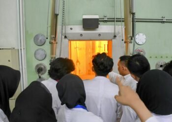 Ruang dengan kaca tebal dan lampu oranye kemungkinan adalah ruang hot cell atau reaktor mini, tempat reaksi nuklir atau penanganan material radioaktif berlangsung secara aman. Dinding dan panel besi tebal merupakan pelindung radiasi.