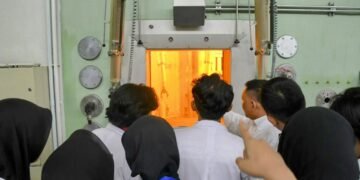 Ruang dengan kaca tebal dan lampu oranye kemungkinan adalah ruang hot cell atau reaktor mini, tempat reaksi nuklir atau penanganan material radioaktif berlangsung secara aman. Dinding dan panel besi tebal merupakan pelindung radiasi.