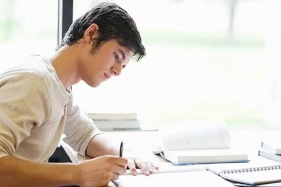 Laki-laki muda itu tampak tekun belajar di meja, menulis sesuatu di buku catatannya sambil dikelilingi tumpukan buku. Cahaya dari jendela membuat suasananya terasa tenang dan fokus.