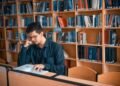 a focused student studying intently amidst the quiet solitude of a library surrounded by shelves of books as they diligently work on an architectural research paper