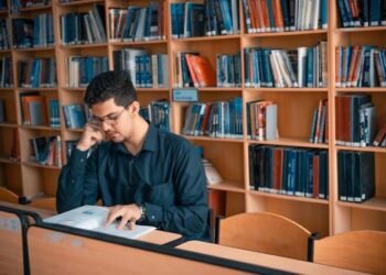 a focused student studying intently amidst the quiet solitude of a library surrounded by shelves of books as they diligently work on an architectural research paper