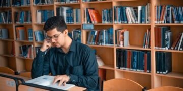 a focused student studying intently amidst the quiet solitude of a library surrounded by shelves of books as they diligently work on an architectural research paper